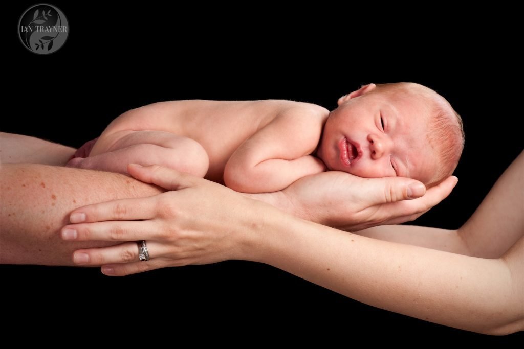 Newborn baby being held in the arms of both parents. Engagement and wedding rings visible.